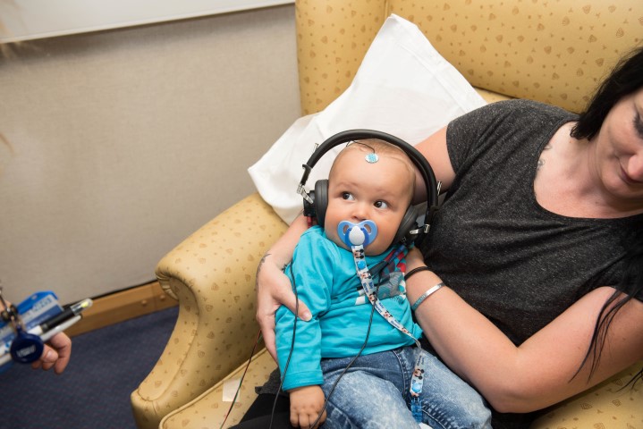 A little baby with a dummy in is wearing headphones while sitting on his mothers lap