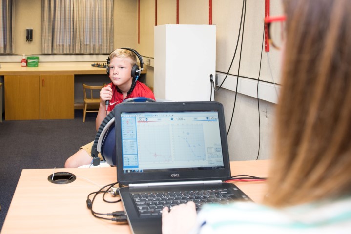 A boy in a red t-shirt listens through headphones during a hearing test. He is holding a stick with a button to push with his thumb. You can see the results of the test on a laptop screen.