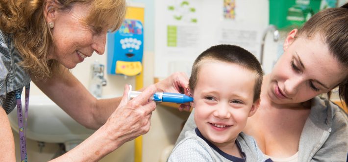 A smiling little boy has a mould made of his ear. He is sitting on his mums knee.