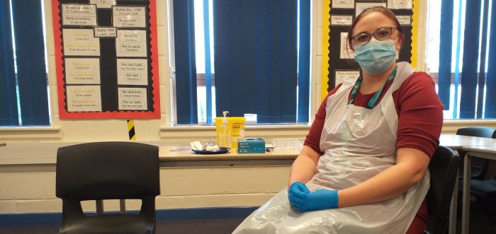 nurse sat in white apron, blue gloves and mask with red top in school classrom