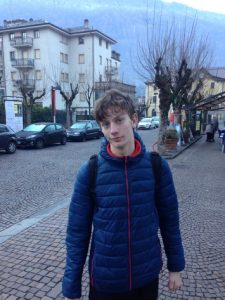 Dan, blue jacket, looking more seriously at the camera, in the background are trees without leaves, a building and parked cars