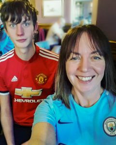 Dan in a red football shirt with his mum in a blue football shirt. its a selfie, both smiling with brown hair