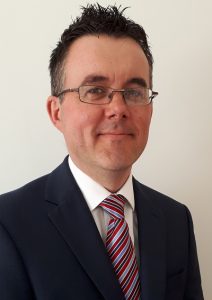 Craig radford in black suit, white shirt and colourful stripey tie smiling at the camera. dark hair and glasses.