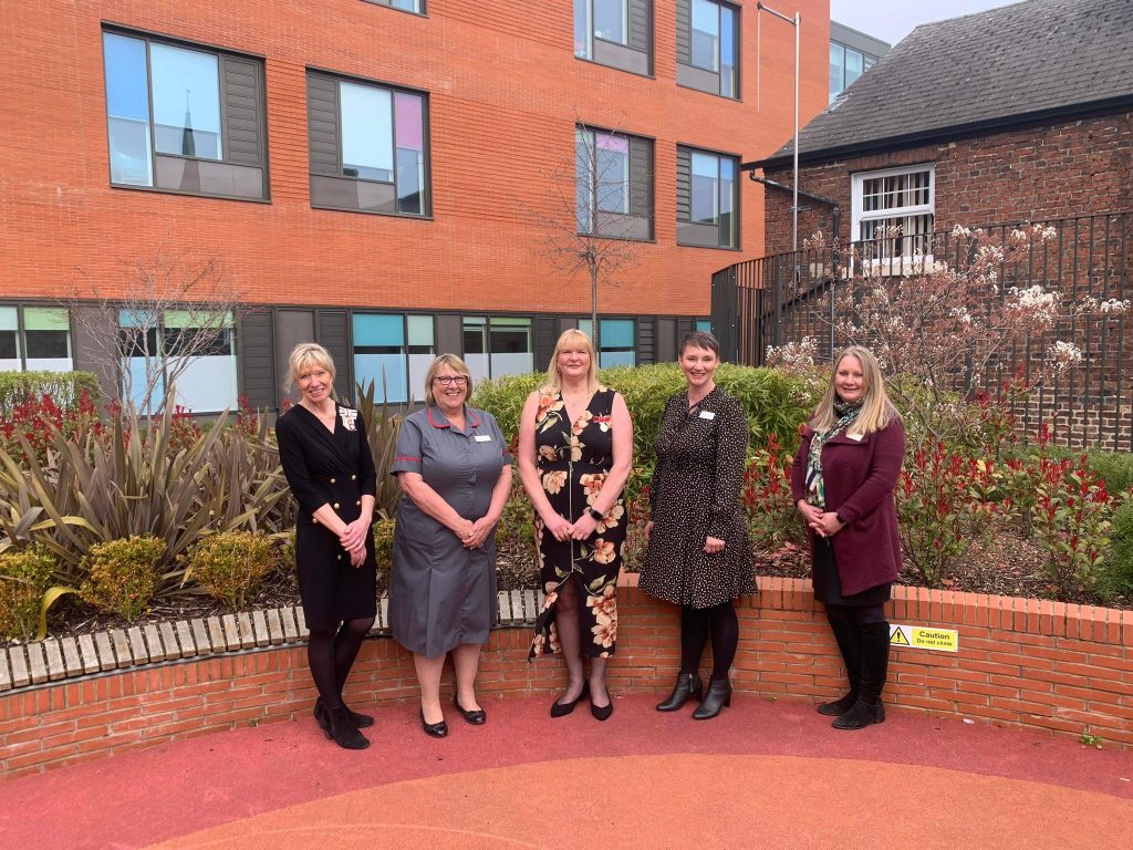 in a row with red brick background and green bushes: Dame Hilary, Director of Nursing Sally Shearer, Nurse Adele Hague BEM, Chief Executive Ruth Brown, Chair Sarah Jones